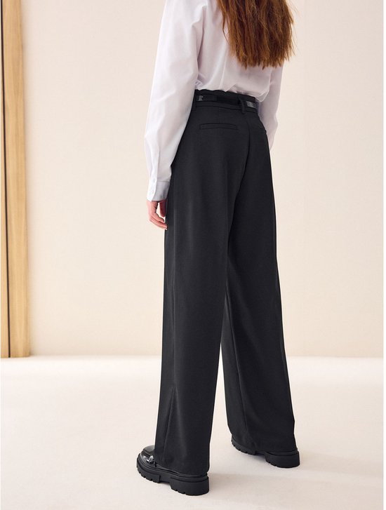 Meisje van achteren gezien, draagt zwarte wijde broek, wit overhemd en zwarte schoenen.