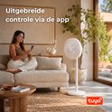 Een vrouw zit op een bank en bedient een witte staande ventilator met haar smartphone. "Uitgebreide controle via de app" staat in tekst op de afbeelding.
