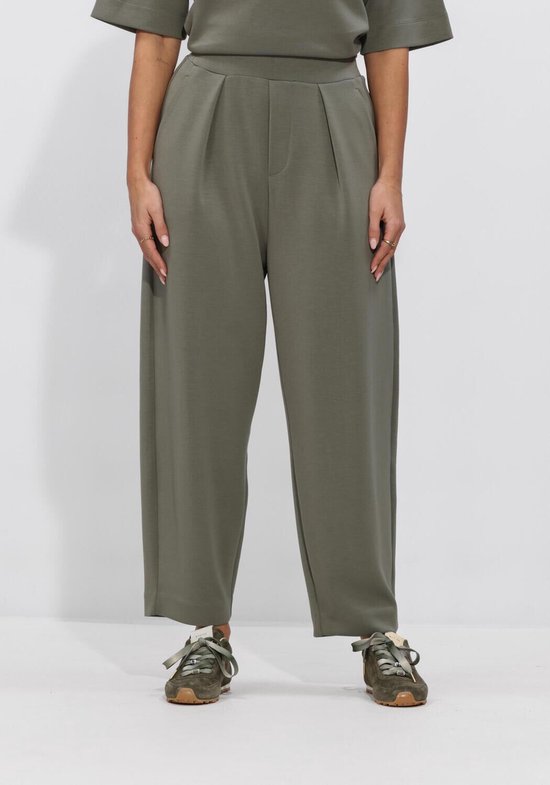 Een vrouw draagt een olijfgroene broek met wijde pijpen, met plooien aan de voorkant. Ze draagt er groene sneakers bij.
