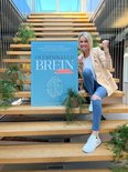 Vrouw met beige blazer, blauwe jeans en witte sneakers zit op een trap naast een boek met de titel 'Overprikkeld brein'.