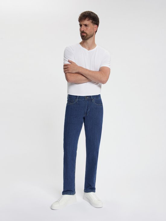 Man met gekruiste armen in donkerblauwe jeans, wit T-shirt en witte sneakers.