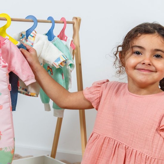 Een meisje glimlacht en pakt een poppenjurk van een kledingrek, met hangers in blauw, geel en roze.