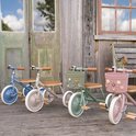 Drie Banwood driewielers op een houten terras: blauw, beige en groen. De groene en roze hebben mandjes met bloemen.