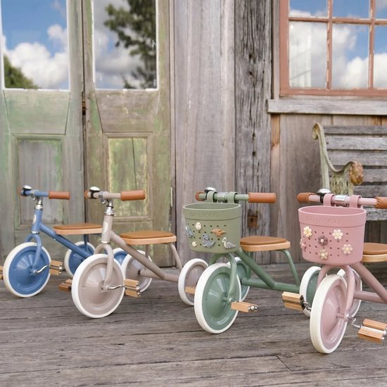 Drie Banwood driewielers op een houten terras: blauw, beige en groen. De groene en roze hebben mandjes met bloemen.