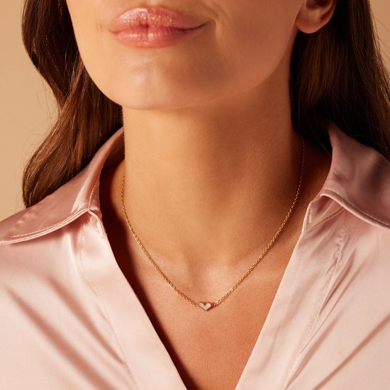 Gouden ketting met een klein wit hartje van parelmoer, gedragen door een vrouw in een roze satijnen blouse.
