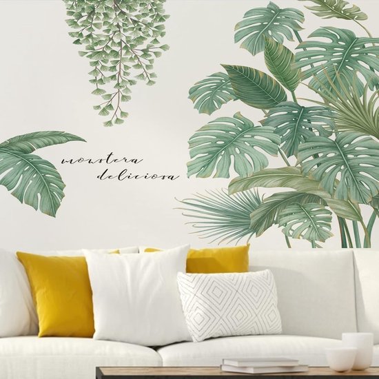 Een muursticker met groene Monstera deliciosa bladeren en hangende varens boven een witte bank met gele en witte kussens.