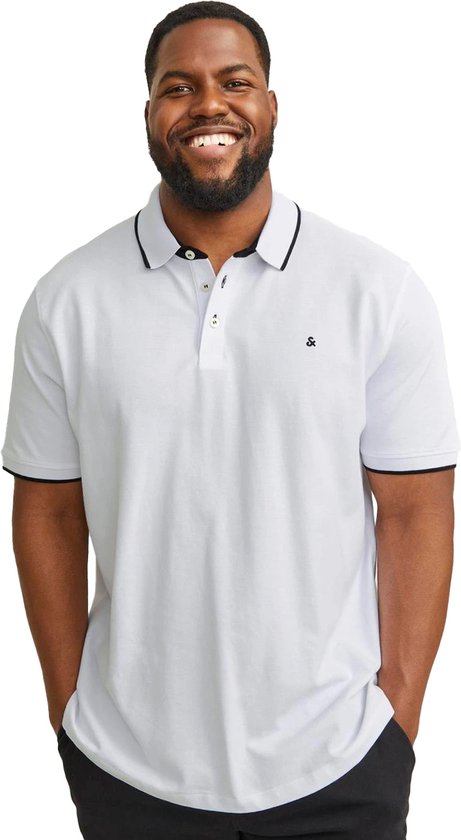 Man in een witte polo met zwarte biezen en zwarte broek. Hij glimlacht met zijn handen in zijn zakken.