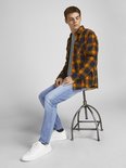 Jonge man in een grijs T-shirt, een oranje met zwarte geruite openvallende blouse en een lichtblauwe jeans.