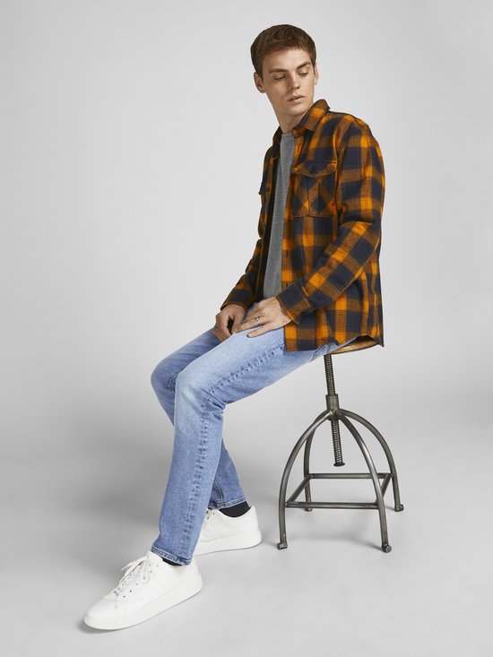 Jonge man in een grijs T-shirt, een oranje met zwarte geruite openvallende blouse en een lichtblauwe jeans.