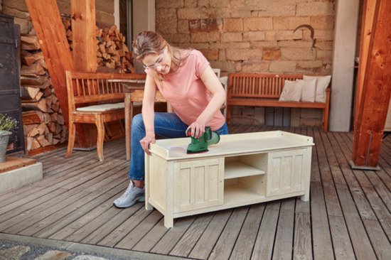 Vrouw schuurt houten kast met groene Bosch multischuurmachine, stofopvangbak bevestigd.