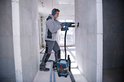 Vakman met gehoorbescherming boort in een muur, verbonden met een blauwe Bosch GAS 18V-10 L stofzuiger.