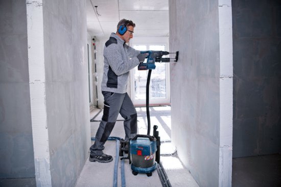 Vakman met gehoorbescherming boort in een muur, verbonden met een blauwe Bosch GAS 18V-10 L stofzuiger.