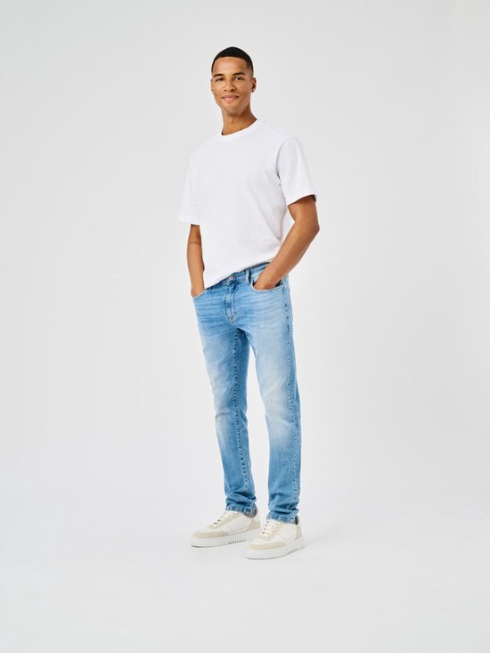 Man met een witte T-shirt en een lichte, gedragen spijkerbroek, staand met handen in zakken.