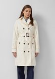 Model draagt crèmekleurige trenchcoat met gestreept overhemd en spijkerbroek. Geknoopte revers.
