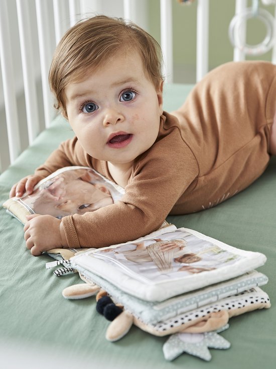 Baby met een bruin rompertje ligt op een zachtgroene ondergrond en bekijkt een stoffen boekje met foto's.