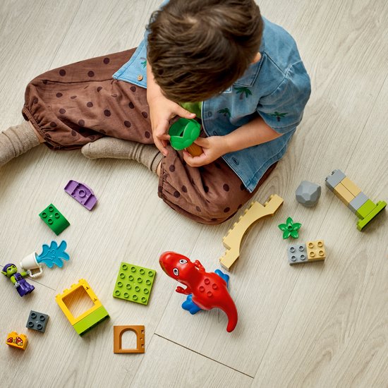 Een jongetje zit op de grond en speelt met losse LEGO Duplo blokken, waaronder een rode dinosaurus en groene goblin.