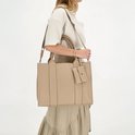Vrouw met beige handtas met 'CHARM LONDON' logo, draagt deze crossbody met schouderband.
