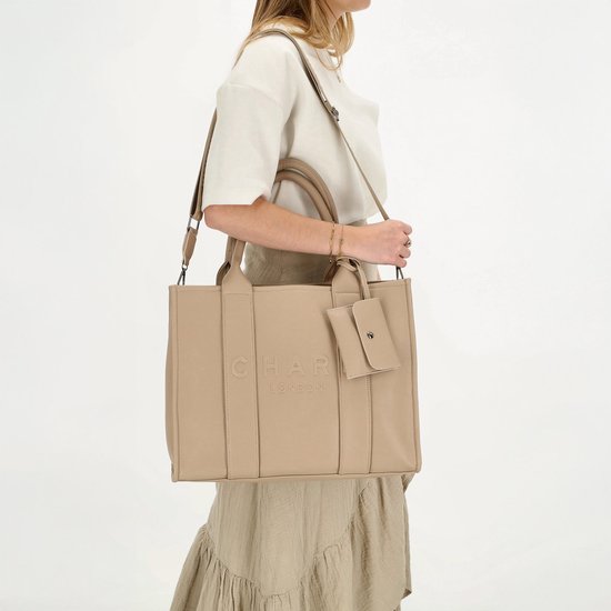Vrouw met beige handtas met 'CHARM LONDON' logo, draagt deze crossbody met schouderband.