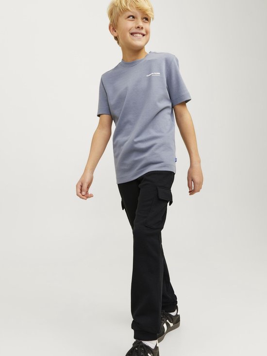 Jongen met blonde haren in een grijze t-shirt en zwarte cargobroek, lachend.