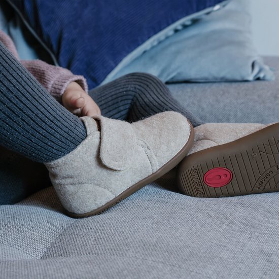 Een meisje op een bank, draagt beige vilten slofjes met klittenband en grijze legging.