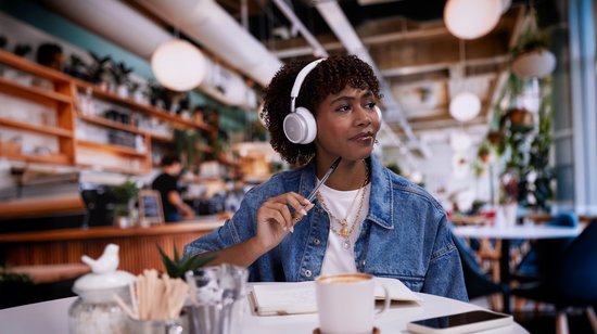 Vrouw met krullend haar draagt witte JBL koptelefoon in een café, zittend aan een tafel.