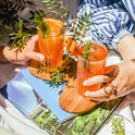 Deux mains tenant des verres de boissons orange, décorés de fruits et d'herbes.