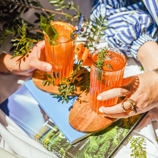 Deux mains tenant des verres de boissons orange, décorés de fruits et d'herbes.