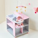 Grijze commode met roze accenten, witte stippen. Een pop met roze slab en muts zit in de kinderstoel. Ernaast liggen gele schoentjes.