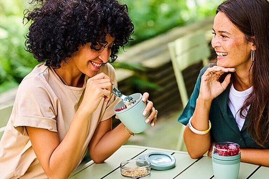 Twee vrouwen zitten buiten te eten uit lichtgroene yoghurtbekers gevuld met muesli en bessen.