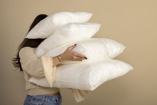 Een vrouw met lange bruine haren draagt een crèmekleurige trui met pofmouwen en een spijkerbroek. Ze houdt een stapel van vier witte kussens vast.