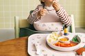 Een baby met een roze slab en rood-wit gestreept shirt eet pasta uit een roze bord op een roze placemat met regenboogopdruk.