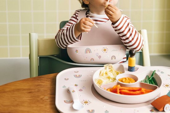 Een baby met een roze slab en rood-wit gestreept shirt eet pasta uit een roze bord op een roze placemat met regenboogopdruk.