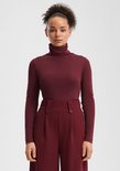 Model draagt een bordeauxrode coltrui en een bordeauxrode broek met hoge taille en knopen.