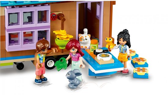 Drie Lego Friends speelfiguren en een grijze konijn bij een opengeklapte Lego tiny house keukentafel.