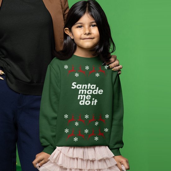 Kind draagt groene kersttrui met rode rendieren, witte sneeuwvlokken en de tekst 'Santa made me do it'.