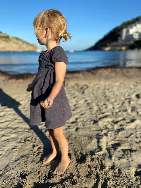 Jong meisje met blond haar in donkerblauw wikkeljurkje op het strand.