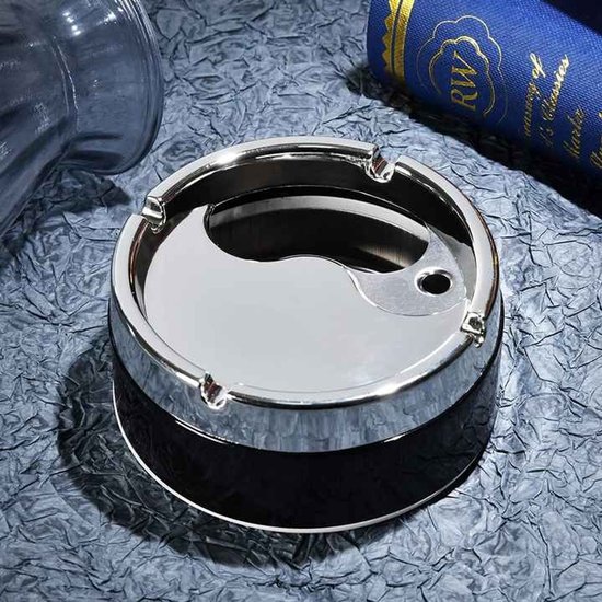 Zilveren draaiasbak met yin-yang motief, chroomkleurige rand en zwarte basis, op een bureau naast een blauw boek.