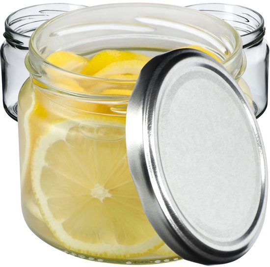Bocal en verre rempli de rondelles de citron, avec un couvercle argenté à côté. Deux bocaux vides à l'arrière-plan.