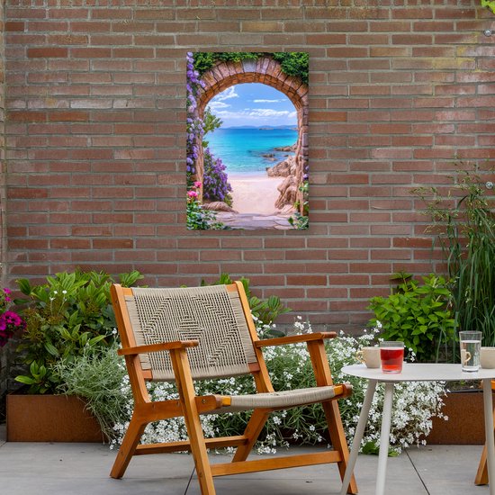Een tuincanvas met een stenen boog en paarse bloemen, die een strand en de turquoise zee toont, gemonteerd aan een bakstenen muur.
