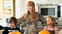 Nicole Kidman met rood haar in een bloemenjurk, staat tussen een jongen en een meisje in een keuken.