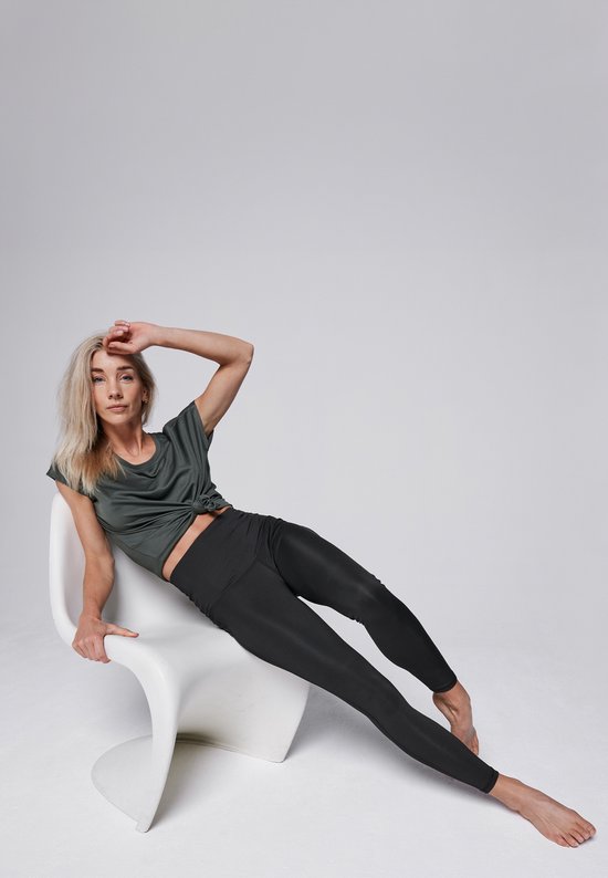 Vrouw zit op een witte stoel, draagt een donkergroen shirt en een zwarte legging, haar hand op haar voorhoofd.