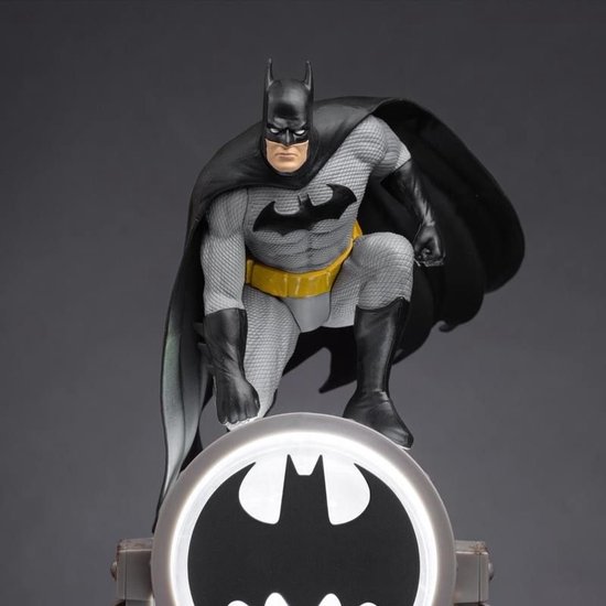 Batman-figuur in grijze outfit hurkend boven de lichtgevende schijnwerper met Bat-logo.