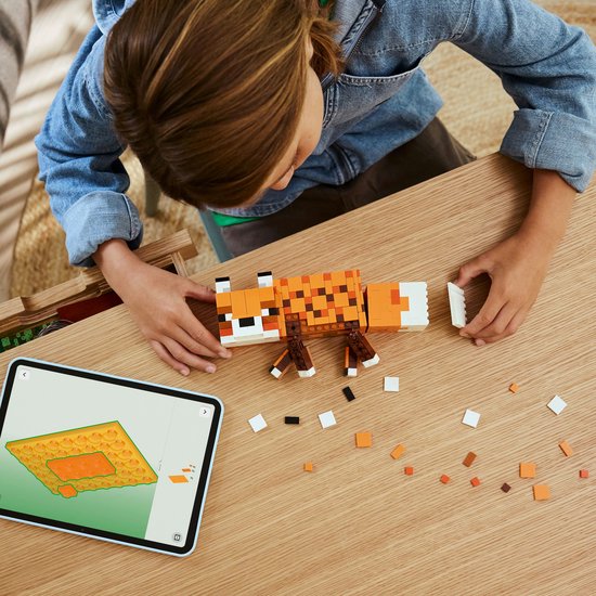 Un garçon en veste en jean et t-shirt vert est assis à une table en bois, construisant avec des pièces LEGO Minecraft et une tablette.