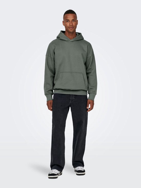 Man van voren, met een groene hoodie en wijde, zwarte spijkerbroek met stonewashed look en sneakers.
