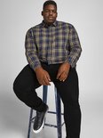Man zittend op een kruk met een geruite blouse en zwarte jeans, met donkere sneakers.