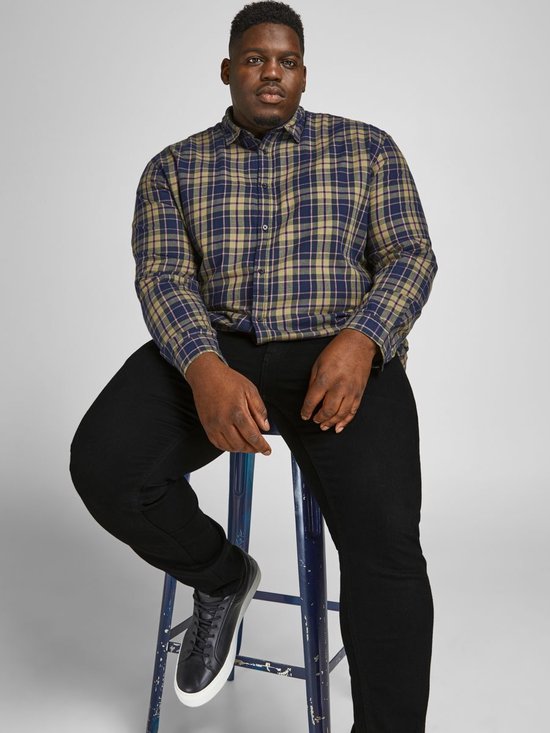 Man zittend op een kruk met een geruite blouse en zwarte jeans, met donkere sneakers.