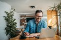 Man glimlacht naar laptop met webcam, schrijft in notitieboekje.