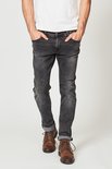 Man met de handen in de zakken van een donkergrijze slim fit jeans met opgerolde pijpen en bruine veterschoenen.