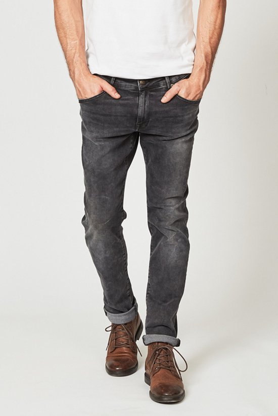 Man met de handen in de zakken van een donkergrijze slim fit jeans met opgerolde pijpen en bruine veterschoenen.