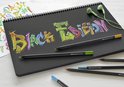 Zwarte schetsboekpagina met "Black Edition" graffiti in felle kleuren, drie zwarte potloden en een groen oortje.